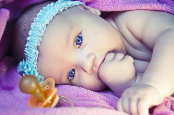 Новорождённая девочка лежит под розовым одеялом рядом с соской и сосёт кулак