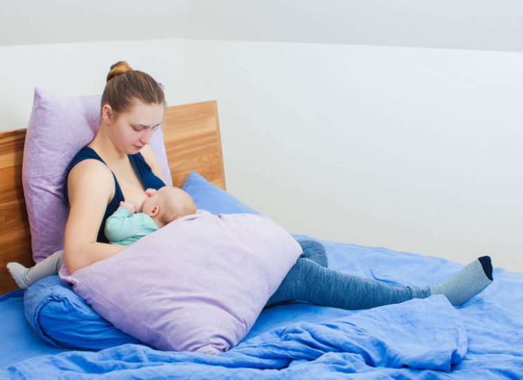 mother sits up on a bed feeding in rugby hold with pillows